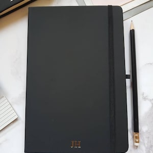 Black A5 notebook with gold personalised initials on the bottom of the journal cover. The notepad has an elastic closure, ribbon page marker and pen loop.