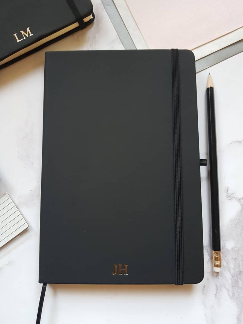 Black A5 notebook with gold personalised initials on the bottom of the journal cover. The notepad has an elastic closure, ribbon page marker and pen loop.