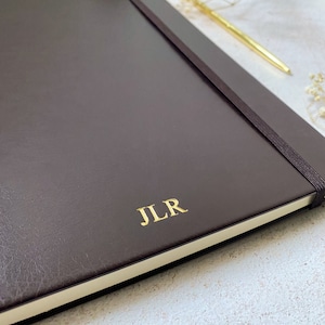 May include: A dark brown leather-bound journal with a gold elastic closure and the initials "JLR" embossed in gold on the front cover.