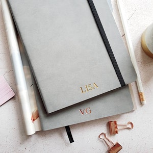 May include: Two gray notebooks with gold lettering, one reading "LISA" and the other "VG." A black elastic band secures the top notebook. A pencil, binder clips, and a magazine are also visible.