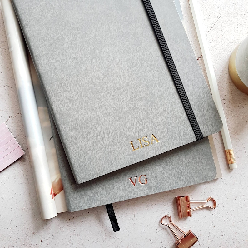 May include: Two gray notebooks with gold lettering, one reading "LISA" and the other "VG." A black elastic band secures the top notebook. A pencil, binder clips, and a magazine are also visible.