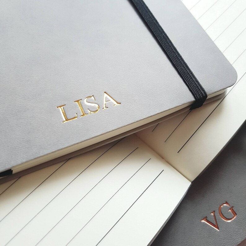 May include: A light gray notebook with the name "LISA" in gold lettering. The notebook has a black elastic band closure. The open notebook reveals lined pages. Another notebook with the initials "VG" is visible.
