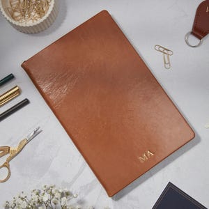 May include: A tan leather journal with the gold initials "MA". The journal is rectangular and lies on a light grey surface, surrounded by stationery items like paperclips, pens, and scissors. A leather keyring is also visible.