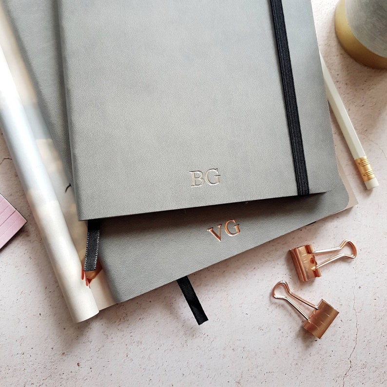 May include: Two gray notebooks with gold-colored initials, a black elastic band, and a black ribbon bookmark. A white pencil, gold-colored binder clips, and a rolled magazine are also visible. The notebooks are on a light pink surface.