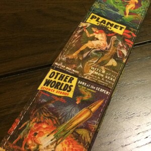 May include: A guitar strap featuring a collage of vintage science fiction comic book covers. The strap displays colorful illustrations and text, including titles like "Amazing," "Weird," and "Other Worlds." The strap is made of fabric and has a black leather end.
