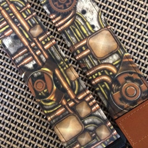 Op de afbeelding: Een gitaarband met een steampunk-ontwerp met koperen pijpen, tandwielen en een textuur achtergrond. De band is bruin met zwarte stiksels en heeft een zwarte verstelbare gesp.