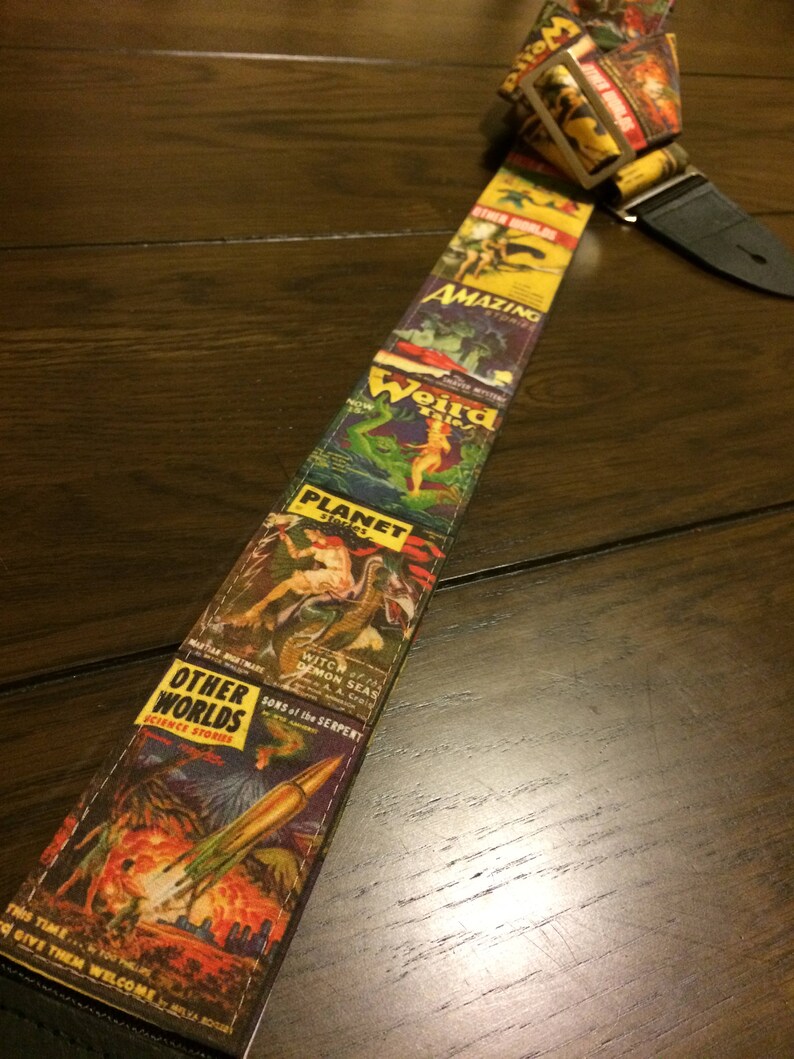 May include: A guitar strap featuring a collage of vintage science fiction comic book covers. The strap displays colorful illustrations and text, including titles like "Amazing," "Weird," and "Other Worlds." The strap is made of fabric and has a black leather end.