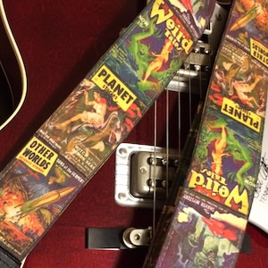 May include: Two guitar straps with vintage science fiction comic book cover designs. The straps feature the titles "Weird Tales," "Planet Stories," and "Other Worlds." The artwork includes images of monsters, rockets, and scantily clad figures. The straps are on a red guitar.