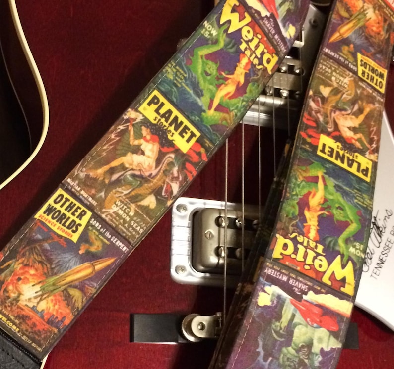 May include: Two guitar straps with vintage science fiction comic book cover designs. The straps feature the titles "Weird Tales," "Planet Stories," and "Other Worlds." The artwork includes images of monsters, rockets, and scantily clad figures. The straps are on a red guitar.
