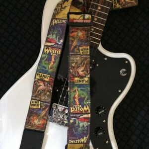May include: A white electric guitar with a black pickguard is shown with two guitar straps. The straps feature colorful vintage comic book covers with the words "Amazing Stories", "Weird Tales", "Planet", and "Other Worlds".