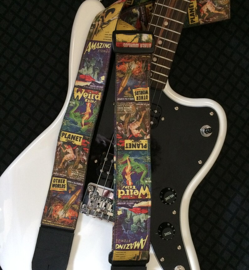 May include: A white electric guitar with a black pickguard is shown with two guitar straps. The straps feature colorful vintage comic book covers with the words "Amazing Stories", "Weird Tales", "Planet", and "Other Worlds".