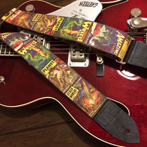 May include: Two guitar straps with a vintage comic book cover design featuring the words "Weird Tales," "Planet Stories," and "Amazing Stories." The straps are black leather with a colorful, retro science fiction theme, resting on a red guitar.