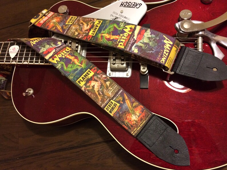 May include: Two guitar straps with a vintage comic book cover design featuring the words "Weird Tales," "Planet Stories," and "Amazing Stories." The straps are black leather with a colorful, retro science fiction theme, resting on a red guitar.