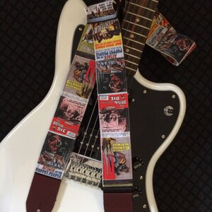 以下が含まれることがあります： 白いエレキギターに2本のギターストラップが付いています。ストラップは、ビンテージのホラー映画のポスターで飾られています。ポスターには、「パペット・ピープルの襲撃」、「死ななかった脳」、「ティーンエイジ・ウェアウルフ」などのタイトルが書かれています。