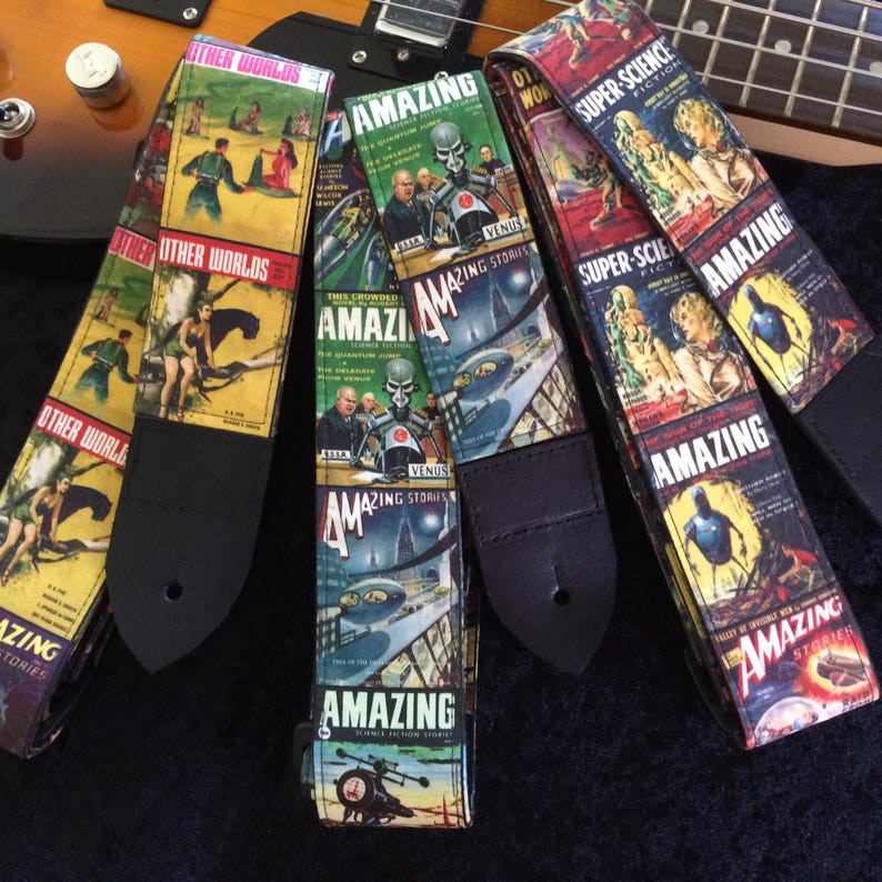 May include: A collection of guitar straps with vintage science fiction book cover designs. The straps feature colorful illustrations and the words "Amazing Stories" and "Other Worlds". The straps are black on the back and have a hole for attaching to a guitar.