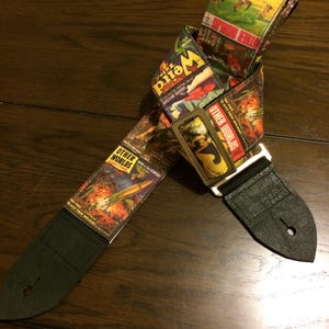 May include: A guitar strap with a vintage comic book design. The strap features images and text from "Weird" and "Other Worlds" comic books. The strap has black leather ends and a metal buckle.