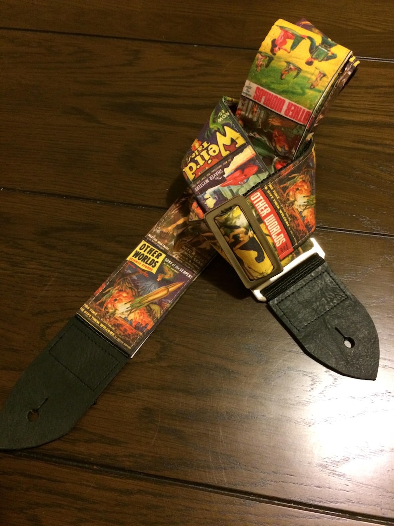 May include: A guitar strap with a vintage comic book design. The strap features images and text from "Weird" and "Other Worlds" comic books. The strap has black leather ends and a metal buckle.