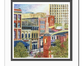 May include: Watercolor painting of a city scene with buildings in shades of red, yellow, and blue. The artwork is 10 inches square and framed in a white mat. The painting depicts a street with people walking.