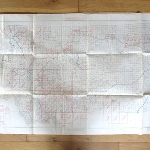 May include: Folded vintage map with red and blue lines, showing geographical features and place names. The map is creased and has a worn appearance, suggesting age and use. The map is unfolded on a wooden surface.