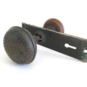 May include: A dark brown, weathered metal door knob set featuring an intricate, embossed geometric and sunburst pattern on the round knob and a rectangular backplate with a central keyhole and decorative etched detailing along the edges.