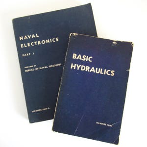 May include: Two vintage navy blue books. The book on the left is titled "NAVAL ELECTRONICS PART 1" and the book on the right is titled "BASIC HYDRAULICS". Both books have white lettering and are prepared by the Bureau of Naval Personnel.