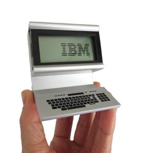 May include: A silver miniature computer with a screen displaying the IBM logo. The keyboard is detailed with small black keys. The device is held in a hand against a white background.
