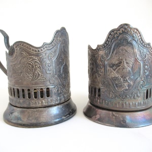 May include: Two vintage silver-toned metal tea glass holders featuring intricate embossed floral scrollwork and a central panel depicting a bird perched on a branch. Each holder has a scalloped upper rim and a wide, flared circular base.