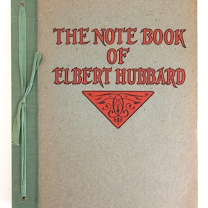 May include: A tan-colored notebook with a green fabric spine and a green ribbon tie. The cover features the red text THE NOTE BOOK OF ELBERT HUBBARD above a red triangular decorative emblem with black filigree details printed on the front.