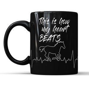 May include: Black ceramic coffee mug with a white design. The design features a running horse silhouette with a heartbeat line and the text "This is how my heart beats".