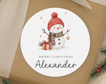 May include: A round white sticker featuring a watercolor illustration of a snowman wearing a red knit hat and a green and red striped scarf. The snowman holds a gold star, stands next to a gift, and the text reads Merry Christmas Alexander.