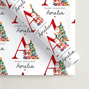 May include: White wrapping paper featuring a repeating pattern of a large red letter A, a watercolor Santa Claus, a decorated Christmas tree, and the text Merry Christmas Amelia in black script. The design includes small red hearts and gold stars.