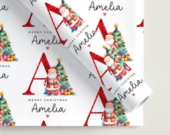 May include: White wrapping paper featuring a repeating pattern of a large red letter A, a watercolour Santa Claus, a decorated Christmas tree, and the text Merry Christmas Amelia in black script. The design includes small red hearts and gold stars.
