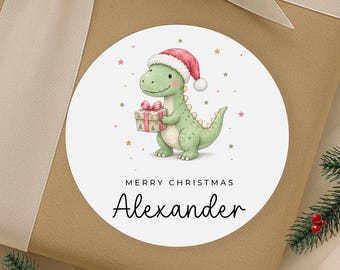 Könnte beinhalten: Ein runder weißer Aufkleber mit einem grünen Cartoon-Dinosaurier, der eine rot-weiße Weihnachtsmütze trägt und ein Geschenk hält. Der Text MERRY CHRISTMAS und der Name Alexander stehen in Schwarz unter dem Dinosaurier, umgeben von Sternen.