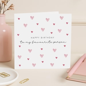 May include: A white birthday card decorated with pink hearts. The card features the message "Happy Birthday to my favourite person" in black script. A pink pen and a pink vase are visible in the background.