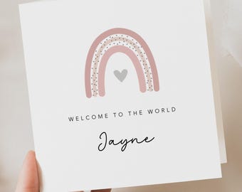 May include: A white greeting card with a pink and gray rainbow design, a gray heart, and the text "WELCOME TO THE WORLD Jayne" in black. The card is held by a hand.