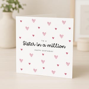 May include: A square white greeting card featuring a pattern of small, light pink watercolor hearts. The text in black reads: TO A Sister in a million HAPPY BIRTHDAY. The card is folded and stands upright, displaying the front cover design clearly.