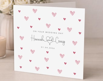 May include: A white wedding card with a pattern of pink hearts. The card reads "On Your Wedding Day" and features the names "Hannah and Craig" with the date "01.04.2026". A bouquet of white roses is in the background.