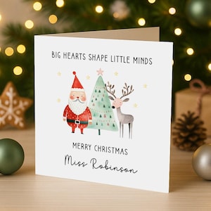 May include: A white square greeting card featuring a watercolour illustration of Santa Claus, a Christmas tree, and a reindeer. The text reads BIG HEARTS SHAPE LITTLE MINDS, MERRY CHRISTMAS, and Miss Robinson in black lettering with small gold stars.