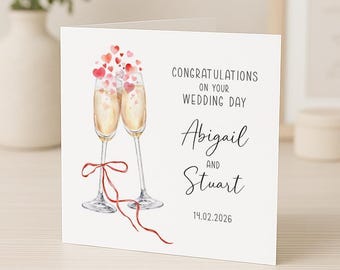 Op de afbeelding: Een vierkante witte wenskaart met een aquarelillustratie van twee champagneglazen verbonden door een rood lint, met zwevende rode en roze hartjes erboven. De tekst luidt: CONGRATULATIONS ON YOUR WEDDING DAY, Abigail AND Stuart, 14.02.2026.