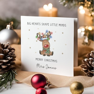 May include: A white square greeting card featuring a cartoon reindeer with a red nose, wearing a green scarf and antlers decorated with colourful Christmas lights. The text reads BIG HEARTS SHAPE LITTLE MINDS, MERRY CHRISTMAS, and Mrs Jones.