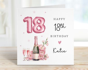 May include: A white greeting card with pink accents, featuring a pink balloon-style "18" and the words "Happy 18th Birthday." A champagne bottle, two glasses, and pink flowers are also depicted. The name "Katie" is written below.