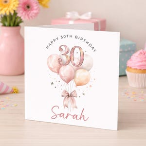 May include: A white square greeting card featuring the text Happy 30th Birthday at the top and the name Sarah in pink script at the bottom. The centre displays a cluster of pale pink and cream balloons tied with a pink ribbon and small stars.
