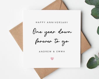 May include: A square white anniversary greeting card featuring the black text HAPPY ANNIVERSARY, One year down forever to go, and the names ANDREW & EMMA, finished with a small pink heart icon, resting on a brown paper envelope.