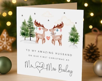 Peut inclure: Une carte de vœux blanche avec une illustration à l'aquarelle de deux rennes ornés de décorations, des sapins verts et des étoiles. Le texte indique : TO MY AMAZING HUSBAND ON OUR FIRST CHRISTMAS AS Mr and Mrs Bailey.