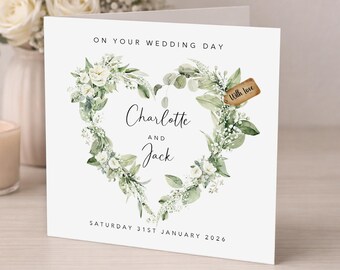 Peut inclure: Une carte de mariage carrée blanche ornée d'une couronne en forme de cœur composée de feuilles d'eucalyptus vertes et de fleurs blanches. Le texte indique ON YOUR WEDDING DAY, Charlotte AND Jack, et SATURDAY 31ST JANUARY 2026, avec une étiquette brune With love.