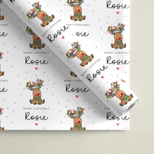 May include: White wrapping paper featuring a repeating pattern of a cartoon reindeer wearing a blue hat and green scarf, decorated with colorful Christmas lights. The text reads Merry Christmas and the name Rosie, accompanied by small red hearts.