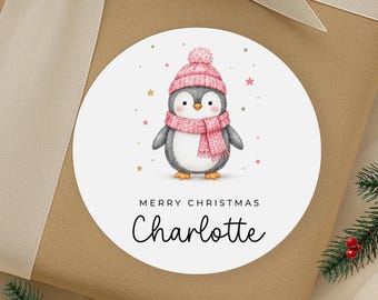 Könnte beinhalten: Ein runder weißer Aufkleber mit einem Cartoon-Pinguin, der eine rosa Strickmütze mit Bommel und einen passenden rosa Schal trägt. Das Design enthält kleine goldene und rosa Sterne. Der Text lautet MERRY CHRISTMAS gefolgt vom Namen Charlotte.