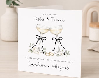 May include: A square greeting card with the text TO A SPECIAL Sister & Fiancée, CONGRATULATIONS ON YOUR ENGAGEMENT, and Caroline ♥ Abigail. The design features two champagne glasses with black ribbons, white flowers, and two gold wedding rings.