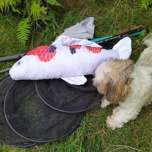 Op de afbeelding: Een witte koi-vis knuffel met rode en zwarte markeringen ligt op groen gras naast een zwart visnet. Een pluizige, lichtbruine en witte hond ligt op het gras.
