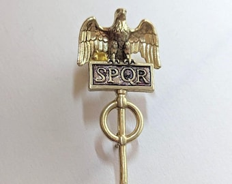 May include: Gold-toned metal pin featuring a detailed eagle perched above a rectangular plaque with the letters "SPQR". The pin has a circular design with a vertical bar extending downwards. The eagle has outstretched wings.